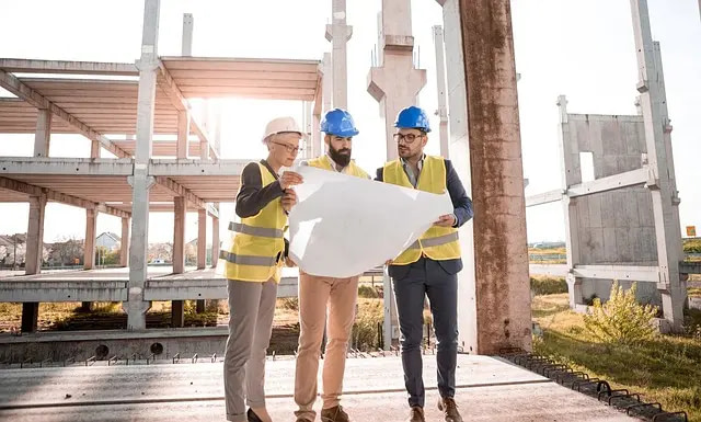 Three Construction Managers Look Over Building Plans At A Construction Site. 