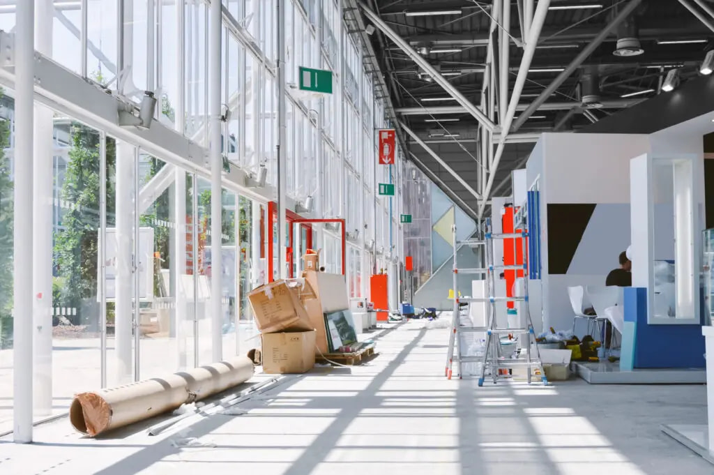 A Busy Exhibition Build Scene With Boxes And Materials On The Floor Adjacent To Two Ladders.
